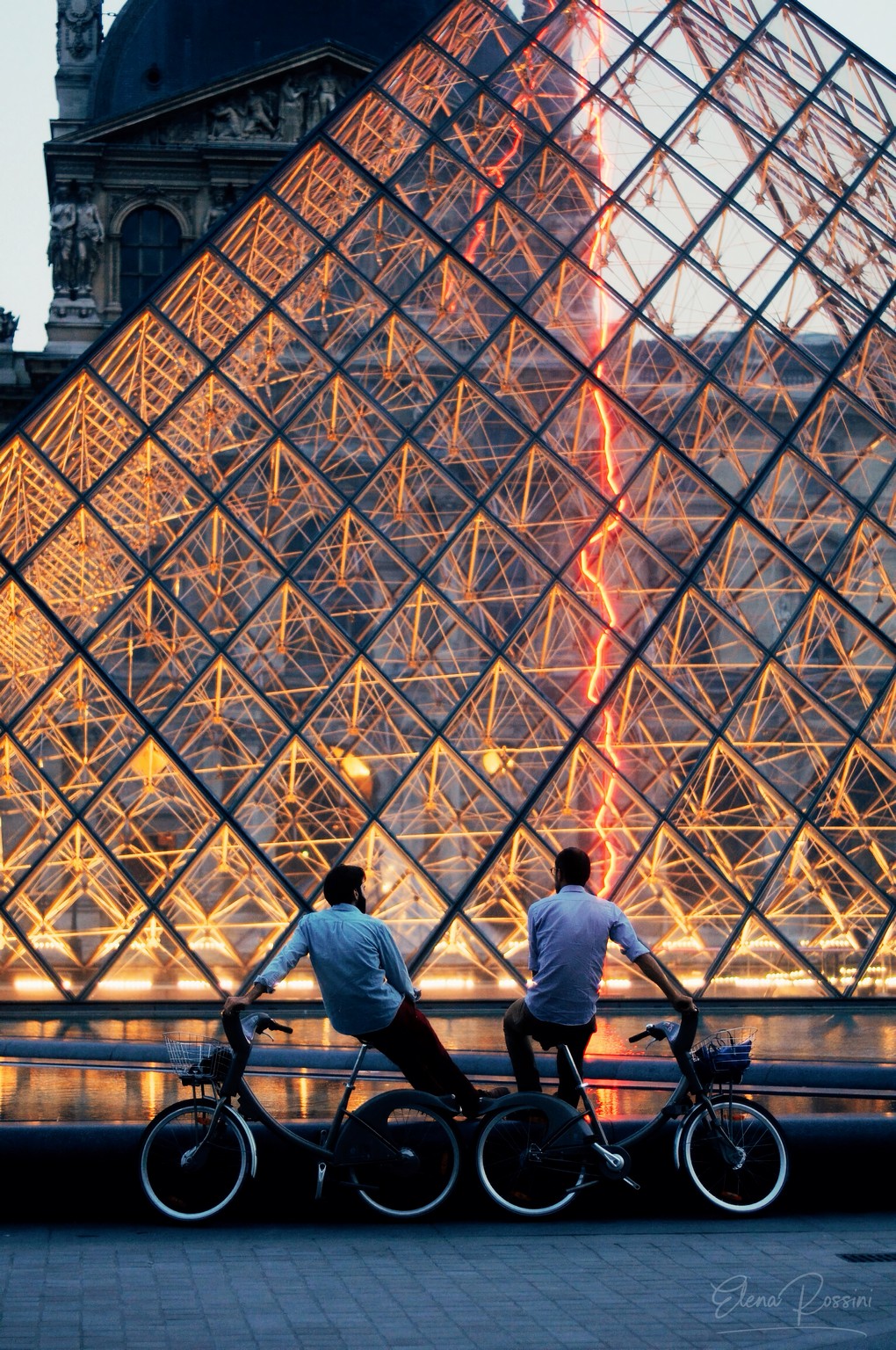 Due amici in bicicletta osservano la piramide del Louvre al tramonto